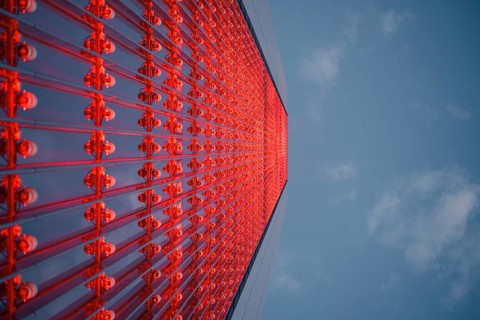 Dramatic upward view of a skyscraper with a red glass facade against a blue sky. The modern