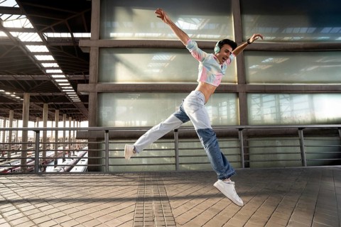 A young person in vibrant attire and headphones joyfully jumps in mid-air on an urban walkway, with