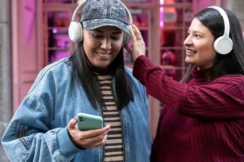 A smiling Latin lesbian couple enjoying music together with headphones. One holds a smartphone,