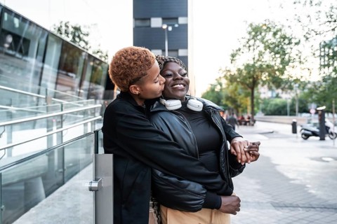 A loving LGBT multiethnic couple is embracing and smiling on an urban street. One partner gently