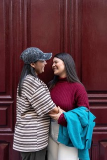 A joyful Latin lesbian couple in a loving embrace stands against a deep burgundy wooden backdrop,