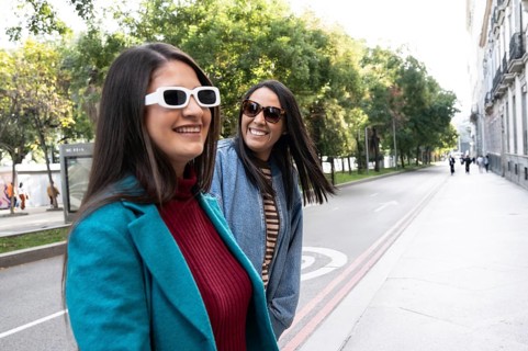 A joyful Latin lesbian couple strolls hand in hand down a tree-lined city street. They wear stylish