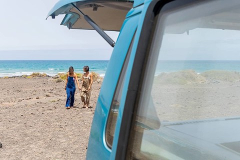 A lesbian couple strolls hand in hand along a beach, adjacent to a classic van. They enjoy the