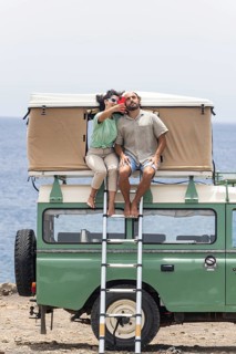A blind man and a woman sit on the rooftop tent of a motorhome, enjoying a coastal view while