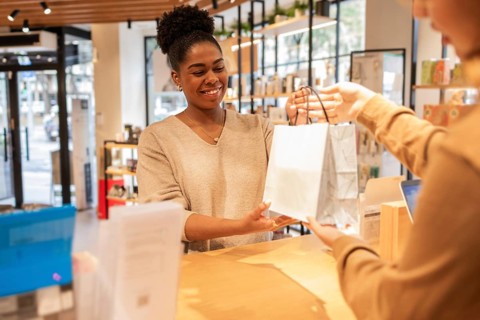 A customer service assistant in a bright cosmetics store smiles while handing a shopping bag to a