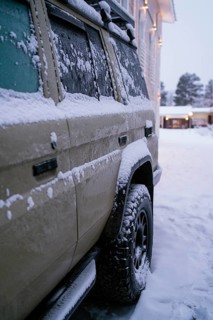 A snow-covered vehicle is parked near a warmly lit building in a snowy setting. The scene captures