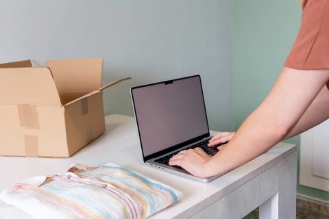 A person uses a laptop to organize and prepare clothing for online sale. The scene includes a