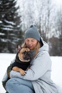 A woman in winter attire lovingly holds her French Bulldog in a snowy landscape. The scene captures