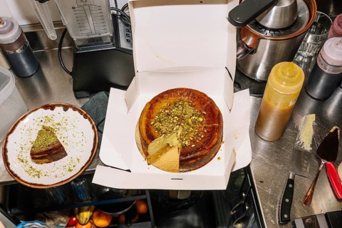 A freshly baked cheesecake topped with crushed pistachios sits in an open box, with a slice plated