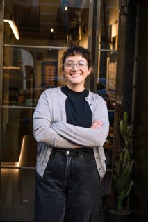 Friendly barista smiling confidently outside a cozy specialty coffee shop. Glass door entrance with