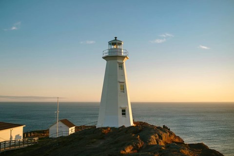 A warm sunset casts a golden glow over the white structure of Cape Spear Lighthouse, perceived as