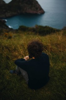 Back view of an unrecognizable man seated on grassy terrain, gazing towards a distant sea with