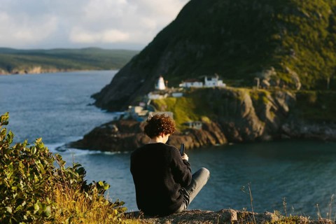 Back view of an unrecognizable man sitting atop a rocky cliff at St. John's, Newfoundland &