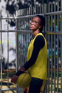 A woman basketball player in a yellow jersey and glasses leans against a metal fence, holding a