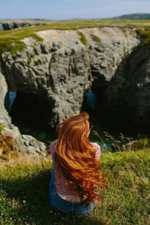 Back view of an unrecognizable redhead woman sitting on a grassy edge, overlooking a rugged coastal