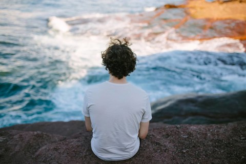 Back view of an unrecognizable person sitting on a rock by the sea, enjoying the serene view at