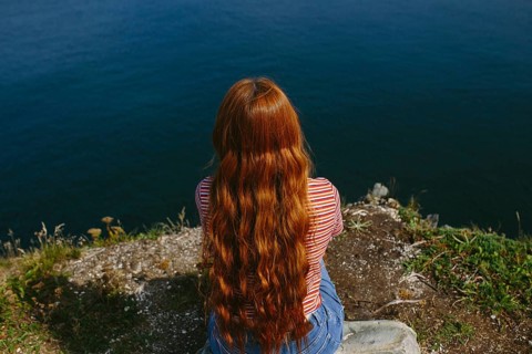 Back view of an unrecognizable redhead female sitting and looking out over the ocean from a cliff.
