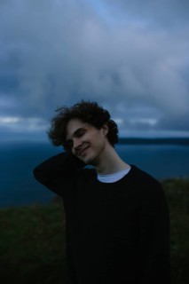 A young man stands with eyes closed relaxed against a backdrop of a cloudy sky and distant sea at
