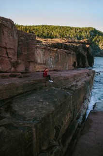 A woman in a red jacket sits pensively on a large rock cliffside, overlooking the ocean at