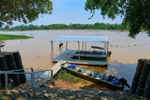 A jetty stretches across the river with a small boat nearby, surrounded by trees, Porto Jofre,