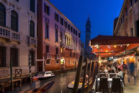 In the evening, lively restaurant on a canal with the tower of Santa Maria della Pietà in the back,