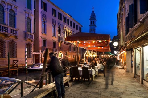 In the evening, lively restaurant on a canal with the tower of Santa Maria della Pietà in the back,