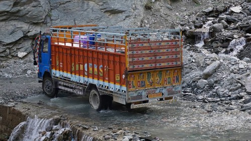 An Indian colorful truck crosses a shallow river on a road surrounded by rocky terrain on the