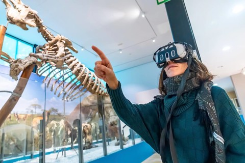 Woman wearing vr headset gestures and points at a life size dinosaur skeleton in a modern natural