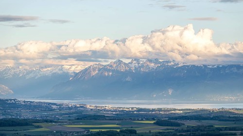 Lake Geneva stretches along the base of towering alpine mountains, with fluffy white clouds casting