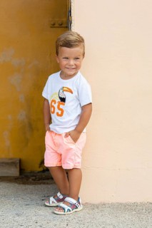 A young boy wearing a white t-shirt with a toucan design and peach shorts leans against a wall He