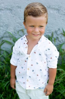 A cheerful young boy with a charming smile stands against a gray wall, dressed in a playful
