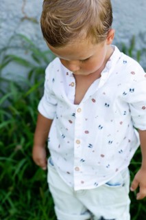 A young boy wearing a white shirt with colorful prints stands amidst green foliage His gaze is