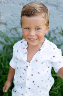 A cheerful young boy in a patterned white shirt smiles brightly at the camera while standing