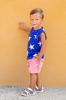 A young child stands against a vibrant orange wall, wearing a blue sleeveless shirt with white