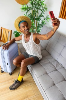 A stylish LGBTQIA+ man prepares for vacation, taking a selfie while holding luggage. The scene