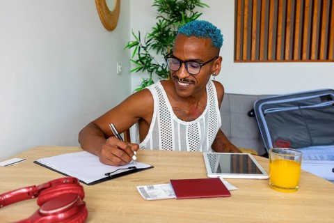 An LGBTQIA+ man enthusiastically prepares for holidays, jotting down plans at a table. Notebooks, a