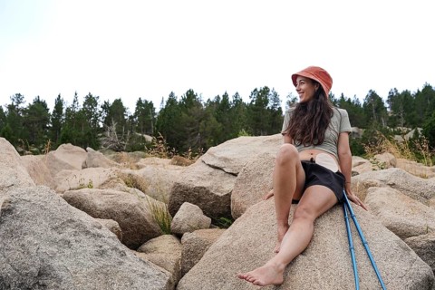 A smiling woman with an ostomy enjoys nature, sitting on large rocks amidst a serene forest