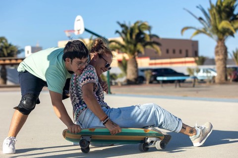 A teenager with Down syndrome helps his mother ride a skateboard outdoors, sharing a fun and