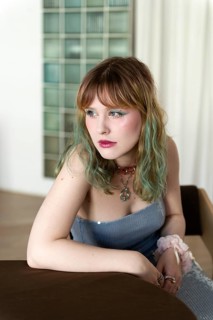 A woman with colorful, pastel hair leans thoughtfully on a brown table Dressed in a light blue