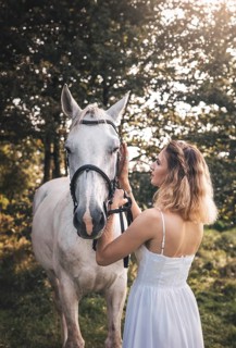 A young woman in a white dress gently caresses a white horse in a sunlit forest. The warm sunlight