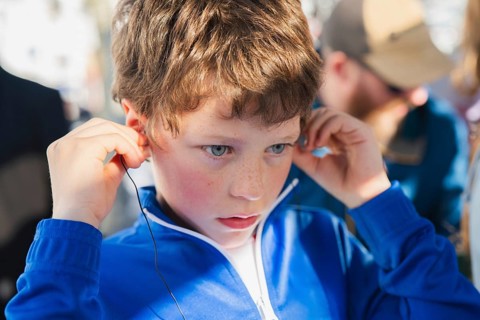 Close-up of a young boy in a blue jacket, adjusting earphones, deeply focused, sitting in a tourist