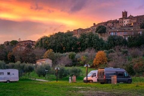 Camper vans and motorhomes parked in a scenic service area below a traditional Tuscany village. The