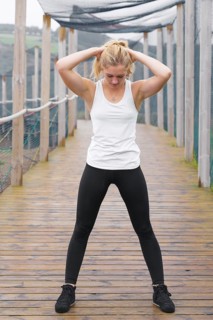A young blond woman exercises by stretching outdoors on a rustic wooden deck, dressed in sporty