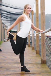 A young blond woman, dressed in athletic wear, stretches on a wooden walkway, displaying a healthy