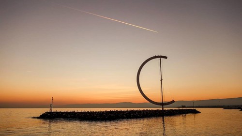 The serene scene of a sunset at Lake Geneva, showcasing a large ring-like sculpture standing on a