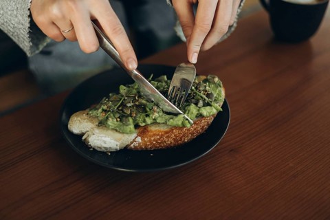 Close-up of hands using a fork and knife to slice avocado toast on a dark plate. The open-faced