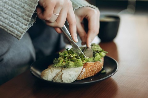 A close-up of hands slicing avocado toast topped with greens on a black plate, accompanied by a