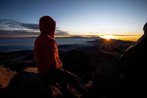 Back view of an unrecognizable man sitting on a mountain peak, watching a stunning sunrise over a
