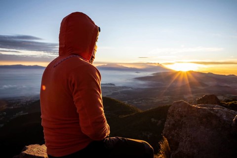 Back view of unrecognizable man sitting atop a mountain, watching the sunrise. The early morning
