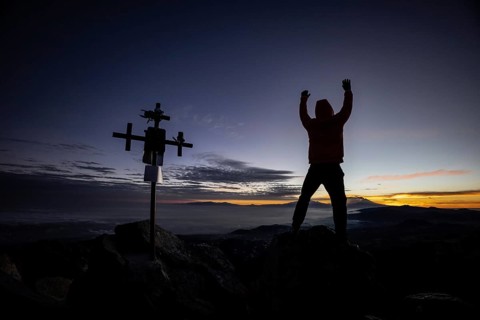 Unrecognizable man stands with arms raised near a silhouetted cross at sunrise, atop a rugged